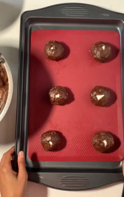 6 dark brown cookie dough balls on baking sheet with silicone mat. small hole of marshmallows peeping out of each dough ball