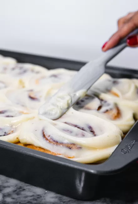 cream cheese icing being spread on freshly baked Homemade Cinnamon Rolls 